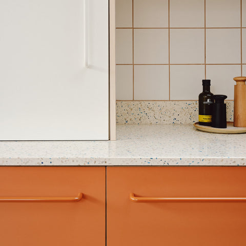 Modern kitchen cabinetry featuring matte orange lower cabinets with matching handles, a white upper cabinet, a speckled quartz countertop, and cream square wall tiles.