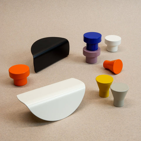 A set of colorful modern hardware pieces and knobs arranged on a textured beige background. The set includes various shapes and colors: a black semicircle, a white semicircle, an orange cylinder with a round top, a purple and blue stacked cylinder, a yellow cone, a gray cone, and another gray cylinder.